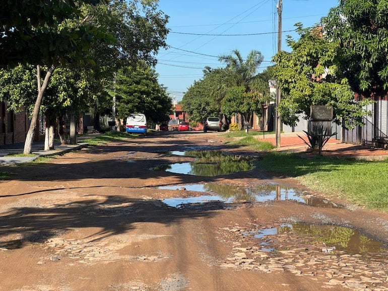 Calle con baches y charcos, rodeada de árboles. Vehículo blanco y azul estacionado a la derecha.