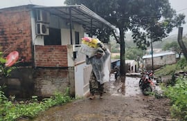 Persona de complexión media en impermeable claro transporta un bulto bajo la lluvia, con una vivienda y una motocicleta de fondo.