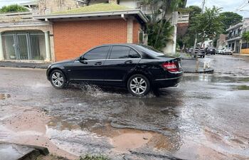 Automóvil negro en movimiento por calle empedrada, salpicando agua en un ambiente urbano nublado.