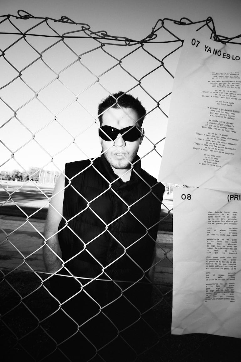 Sebastián González tras malla metálica, con gafas oscuras y chaleco negro, rodeado de un ambiente abierto y dramatismo en blanco y negro.