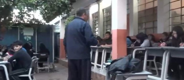 Grupo de estudiantes en ropa oscura, tomando apuntes, mientras un docente de espaldas explica frente a ellos en un patio escolar.