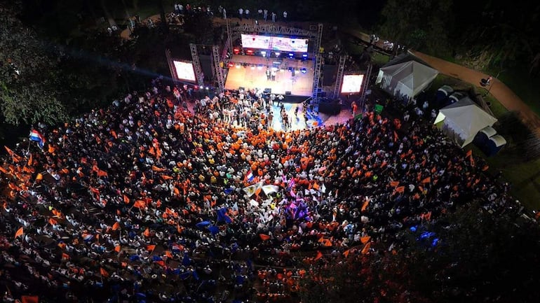 Multitud animada con banderas naranjas en evento al aire libre, con oradores destacados en el escenario y carpas alrededor.