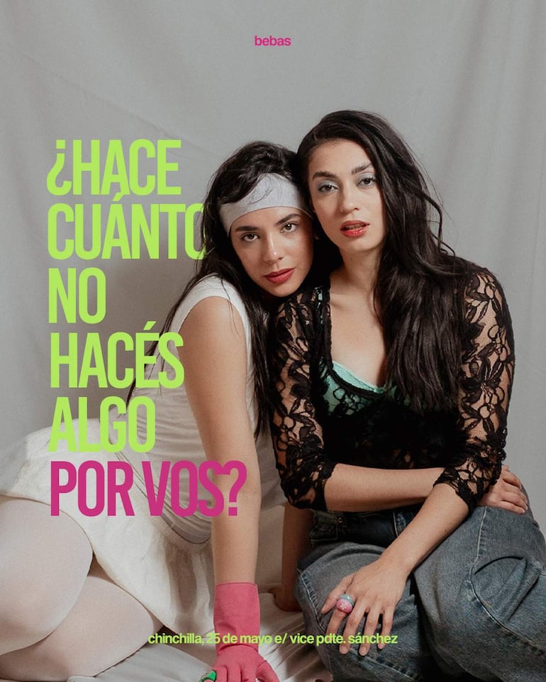 Dos mujeres, una con vestido blanco y otra en blusa negra, lucen relajadas en un ambiente artístico, mirando a la cámara.