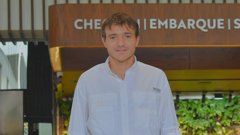 Hombre en camisa blanca de manga larga frente a cartel de embarque en ambiente de aeropuerto.