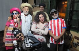 Seis actores sonríen en disfraces coloridos, incluyendo sombreros y gafas, en un ambiente festivo y acogedor.