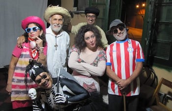 Seis actores sonríen en disfraces coloridos, incluyendo sombreros y gafas, en un ambiente festivo y acogedor.