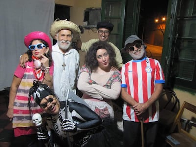 Seis actores sonríen en disfraces coloridos, incluyendo sombreros y gafas, en un ambiente festivo y acogedor.