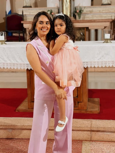 Mujer con cabello oscuro y vestido rosa claro sonríe mientras sostiene a una niña con vestido y diadema rosa en una iglesia decorada.