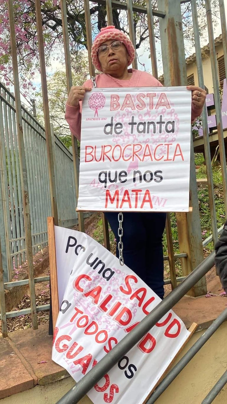 Mujer de pie con suéter rosa y gorro, sostiene un cartel de protesta sobre burocracia y salud.