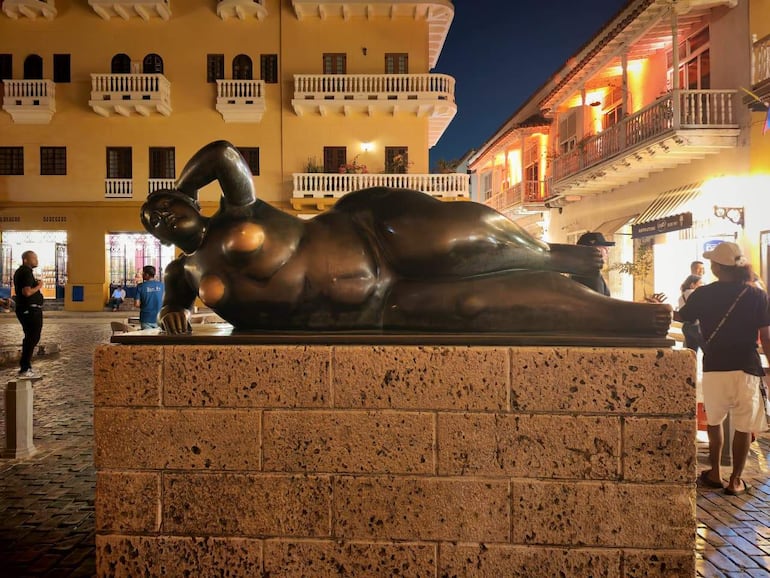 Transeúntes contemplativos en plaza iluminada, con escultura de figura femenina y edificios históricos de fondo.