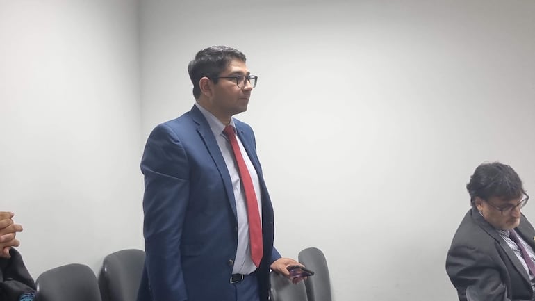 Hombre en traje azul y corbata roja de pie, mientras otro en traje negro lo escucha atentamente en un ambiente interior.