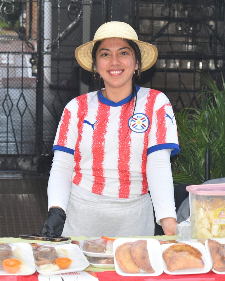 Mujer con camiseta del equipo paraguayo sonríe detrás de una mesa con empanadas en un ambiente festivo.