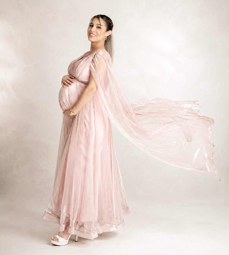 Mujer embarazada sonriente en vestido rosa claro y capa transparente, sostiene su vientre en un entorno suave y sereno.