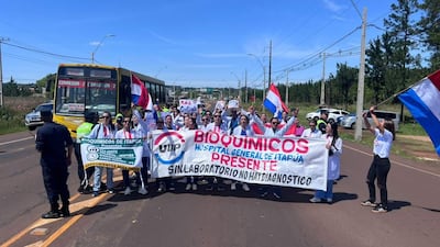 Grupo de bioquímicos en batas blancas sostiene pancartas en una carretera, protestando con banderas paraguayas de fondo.