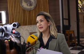 Mónica Recalde en chaqueta verde pálido y blusa negra, sostiene documentos y un micrófono amarillo en un ambiente elegante.
