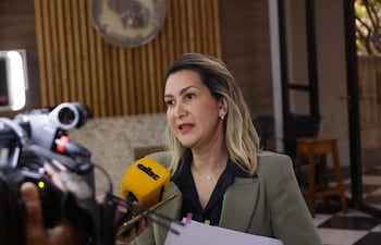 Mónica Recalde en chaqueta verde pálido y blusa negra, sostiene documentos y un micrófono amarillo en un ambiente elegante.