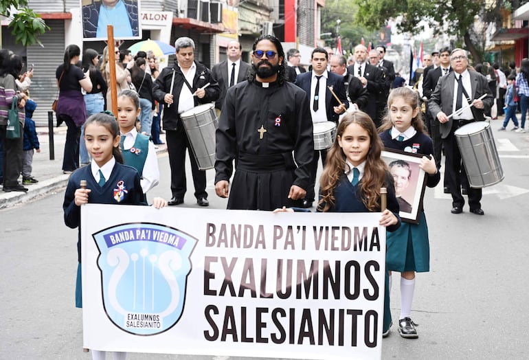 Dos niñas con uniformes escolares sostienen un banner, mientras un adulto muestra un crucifijo en un entorno urbano festivo.