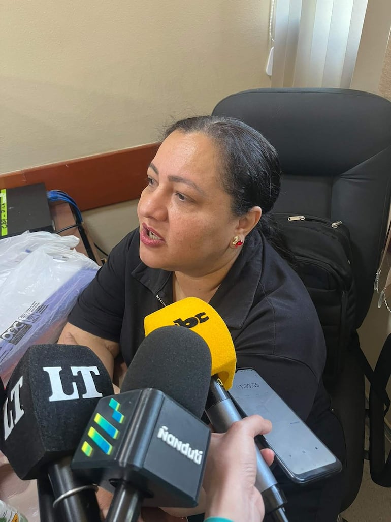 Sergia Encina, mujer de cabello oscuro y camiseta negra, sentada en una oficina, habla ante varios micrófonos de medios.