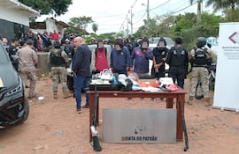 Cuatro detenidos con capuchas en la cabeza, parados detrás de una mesa donde se exhibe parte de lo incautado en el procedimiento