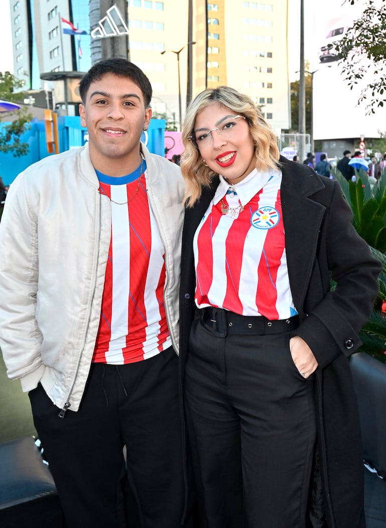Jóvenes sonrientes, uno con camisa de la APF y el otro con gafas, en ambiente festivo al aire libre.