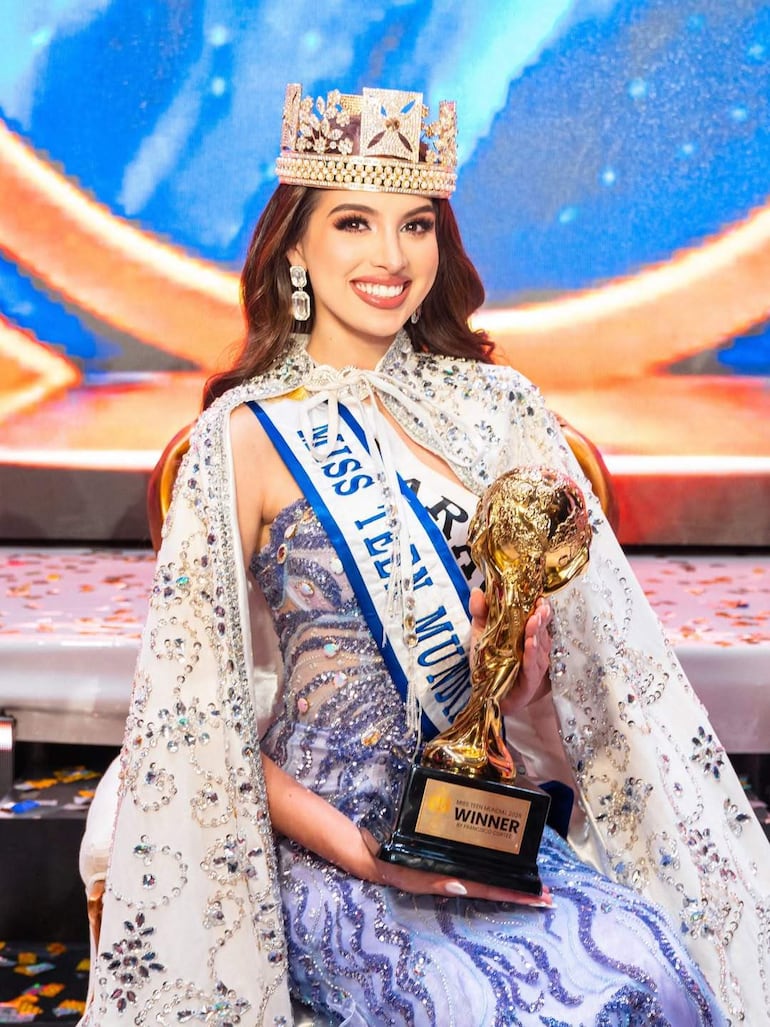 Refrancisco Cortez sonríe con una corona y trofeo dorado, vistiendo un brillante vestido y banda de Miss Teen Paraguay, en un ambiente festivo.