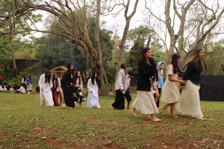 Diez mujeres jóvenes en vestidos claros caminan organizadas por un espacio verde, rodeadas de árboles y un ambiente festivo.