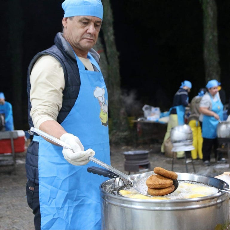 Hombre de mediana edad con delantal y gorro de chef, sostiene una paleta con comida frita en exterior, rodeado de personas con delantales azules.