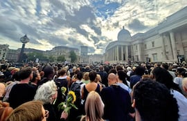 Personas de pie en Trafalgar Square, observando un evento, algunas sosteniendo girasoles, con cielo nublado de fondo.
