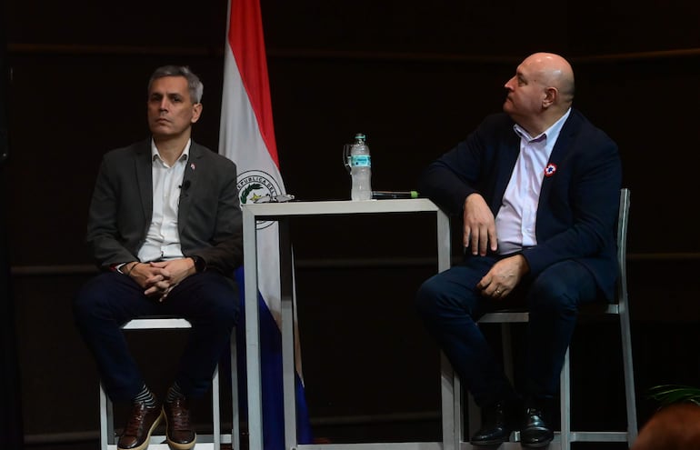 Hombre canoso en blazer oscuro y camisa blanca escucha atentamente a compañero de cabeza rapada, ambos en ambiente formal con botella de agua entre ellos.