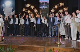 Grupo de personas en trajes formales levantando copas, celebrando un logro en un escenario con luces brillantes y fondo oscuro.