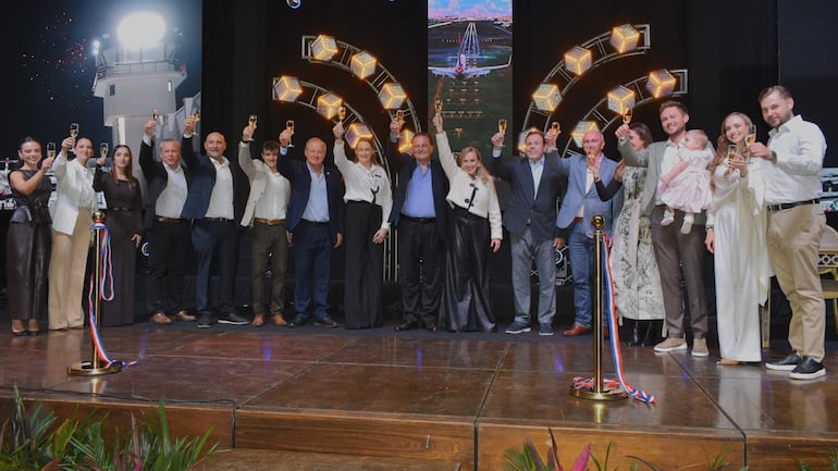 Grupo de personas en trajes formales levantando copas, celebrando un logro en un escenario con luces brillantes y fondo oscuro.