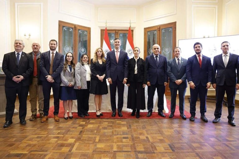 Grupo de doce personas posando en un elegante interior, con un hombre en traje claro al centro y mujeres y hombres a su alrededor.