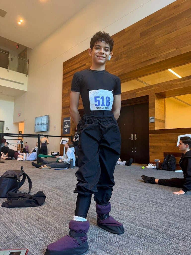 Joven sonriente de pie con camiseta negra y pantalones deportivos, auriculares, rodeado de personas sentadas en un ambiente luminoso.