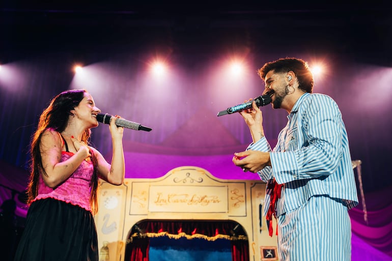Mujer con blusa rosa y falda negra, hombre en pijama de rayas, cantan en un escenario vibrante.