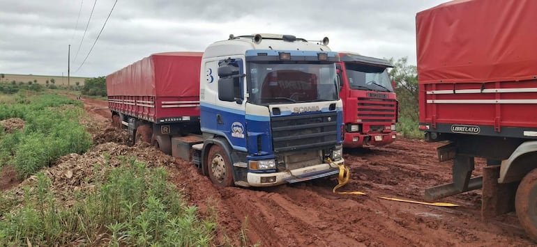 Camiones Scania azul y rojo estancados en barro en un camino rural, sin personas visibles y cielo nublado.