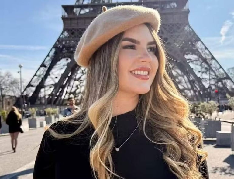 Mujer con cabello largo y boina clara sonríe en un día soleado, con la Torre Eiffel al fondo.