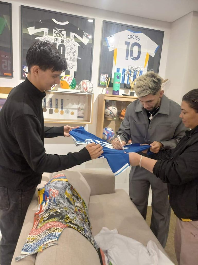 Julio Enciso en traje gris firmando camiseta azul, mientras Ramón Sosa, en suéter negro, lo escucha atentamente en entorno deportivo.