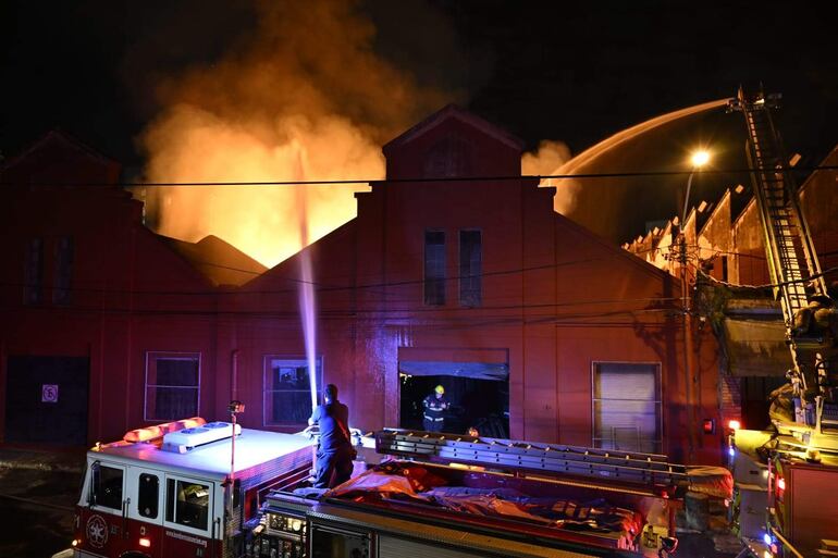 Dos bomberos en acción, uno en camión dirigiendo una manguera y otro en la entrada del edificio en llamas.