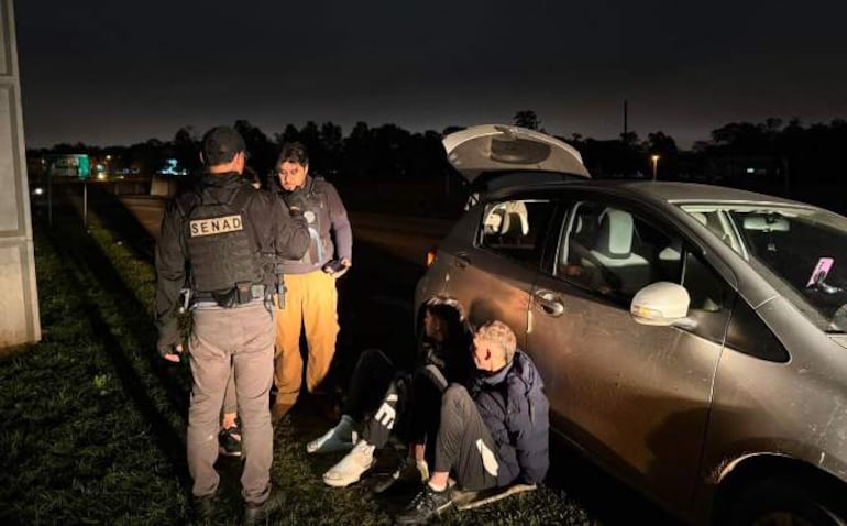 Dos jóvenes sentados con ropa oscura, un agente de SENAD de pie frente a ellos, un auto compacto gris con el maletero abierto.