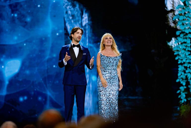 Hombre en traje azul gesticulando en el escenario, mientras una mujer con vestido largo azul sonriendo lo acompaña. Público visible en el fondo.