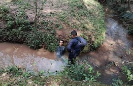 Hombre en agua marrón con camiseta oscura y cabello corto es observado por otra persona de pie en la orilla con traje oscuro.