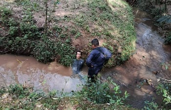 Hombre en agua marrón con camiseta oscura y cabello corto es observado por otra persona de pie en la orilla con traje oscuro.