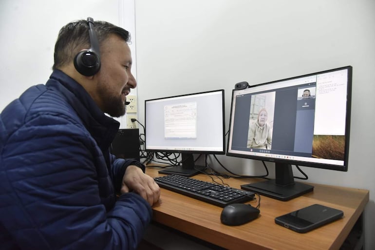 Derlis León, sentado frente a la computadora con chaqueta azul y auriculares, sonríe mientras conversa por videoconferencia con una mujer en la pantalla.