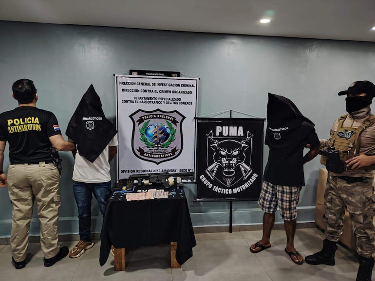 Dos hombres con capuchas, uno en camiseta blanca y otro en negra, son sostenidos por un policía con uniforme antinarcóticos.