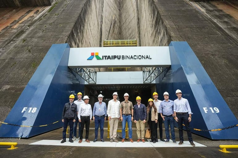 Christopher Landau, Javier Giménez y otros visitantes en cascos de seguridad, sonriendo frente a la entrada de Itaipú.