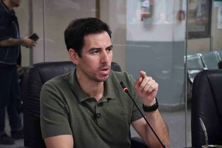 Carlos Reyes, con camiseta verde y gesto serio, conversa en una reunión; un hombre de pie lo acompaña parcialmente visible.