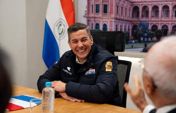 Santiago Peña sonriente en chaqueta oscura, con emblema, en mesa con botella de agua y tapete de la bandera paraguaya.