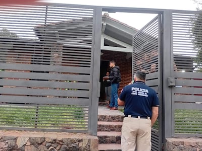 Hombre en abrigo negro sostiene documentos mientras conversan una mujer vestida casual y un policía de espaldas con gorra en un ambiente urbano.