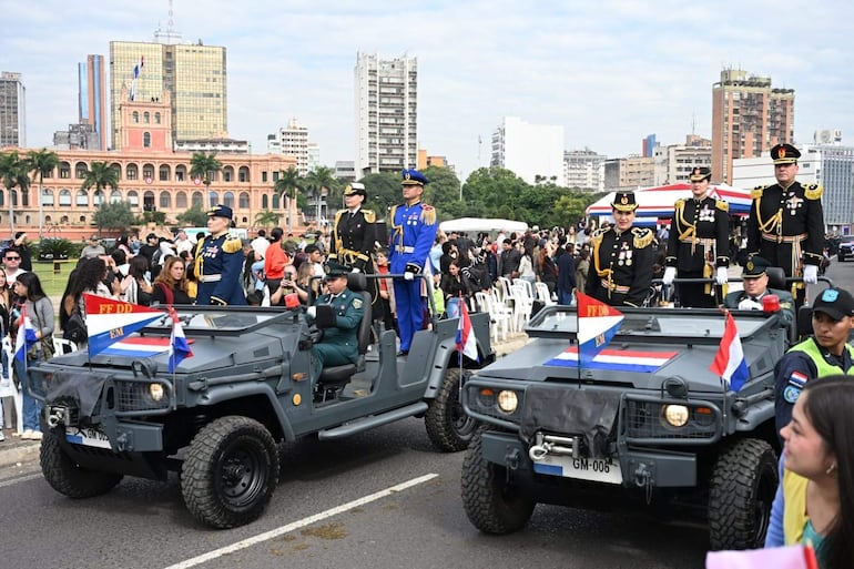 Militares en uniforme formal desfilan sobre vehículos, rodeados de público en sillas, frente a un emblemático edificio.