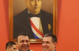 Miguel del Puerto con chaqueta clara y negra, ríe abrazando a Horacio Cartes, en traje oscuro y corbata roja, en ambiente institucional.